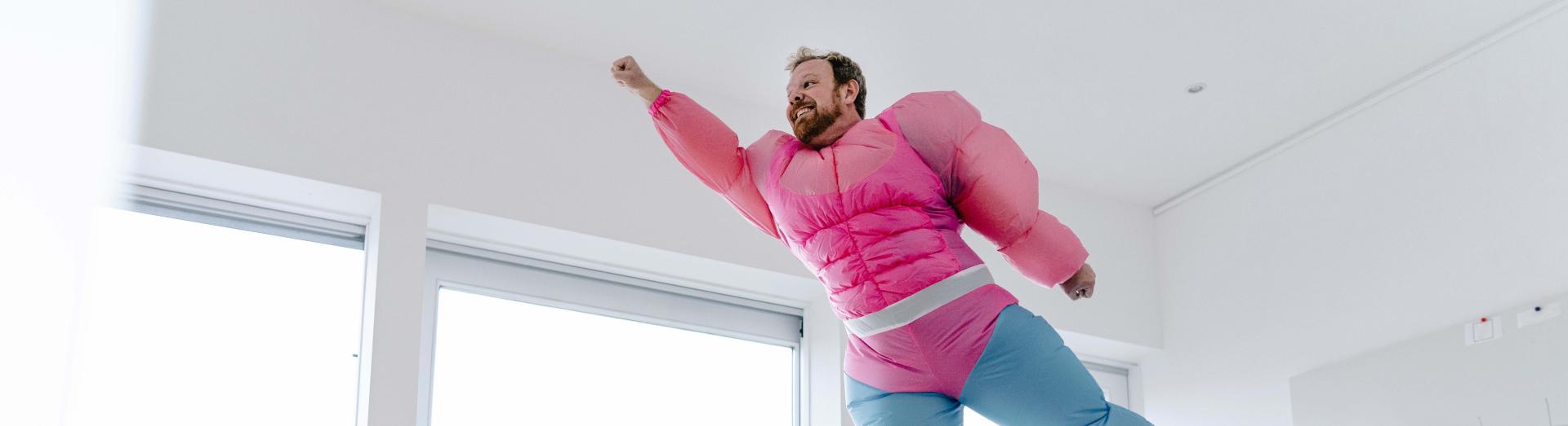 Mann in rosa Michelin-Männchen steht bloßfüssig am Tisch und streckt Arm und Bein weg