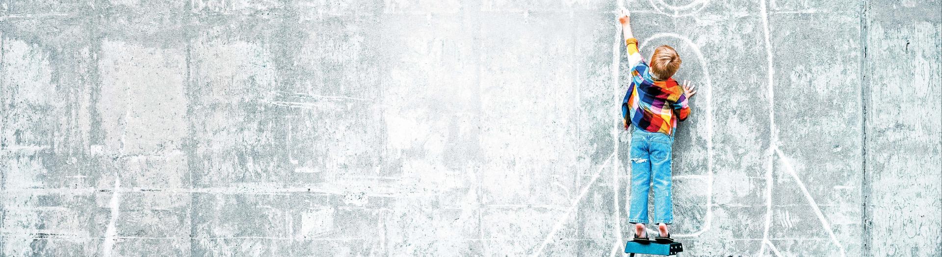 Kleiner Bub auf dreistufiger Leiter zeichnet mit Kreide Rakete an eine Wand