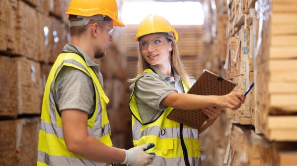 Zwei Personen mit gelben Bauhelm und Warnweste in einer Holz-Lagerhalle
