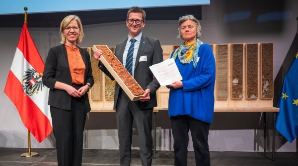 Preisverleihung an die DGK Krems - drei Personen mit Fahnen im HIntergrund