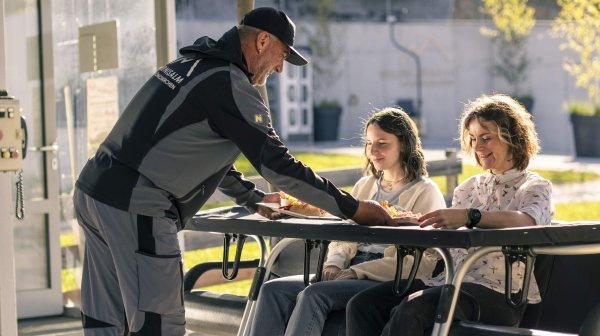 zwei Personen auf einem Sessellift und eine Person stellt etwas auf den Sessellift