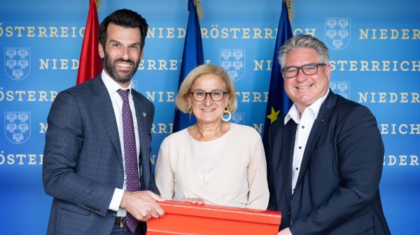 Drei Personen halten ein Stück eines roten Gleisabschnittes vor einer Niederösterreich-Rückwand in die Kamera