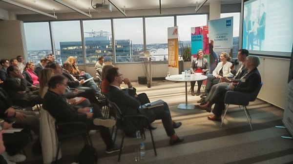 Raum mit 4 Vortragenden, Personen mit Kinobestuhlung, Budapest im Fenster dahinter zu sehen