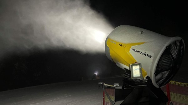 Schneekanone bläst Schnee auf eine präparierte Piste bei Nacht. Das Gerät ist gelb-weiß und steht neben einem roten Sicherheitsnetz, während im Hintergrund eine verschneite Landschaft und eine Lichtquelle sichtbar sind.