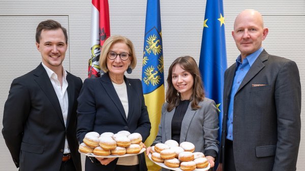 Vier Personen stehen vor einer österreichischen, einer niederösterreichischen und einer EU-Fahne und halten mehrere Tabletts mit Krapfen.