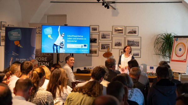 Ein voller Workshop‑Raum, in dem eine Vortragende vor einer Gruppe von Teilnehmenden steht. Auf einem großen Bildschirm ist die Präsentation mit dem Titel ‚Zukunftsworkshop – Leben und Arbeiten im Weinviertel‘ zu sehen.