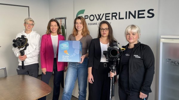 Eine Gruppe von fünf Personen steht in einem Büroraum vor einer Wand mit dem Logo ‚Powerlines – An Equans Company‘. Zwei Personen halten professionelle Kameras, während eine andere Person einen Würfel mit dem Aufdruck ‚LEADER Region Weinviertel Ost‘ präsentiert.