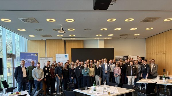 Große Gruppe von Menschen steht in einem modernen Konferenzraum für ein Gruppenfoto zusammen. Im Vordergrund befinden sich mehrere Tische mit Unterlagen, Getränkeflaschen und Stühlen.