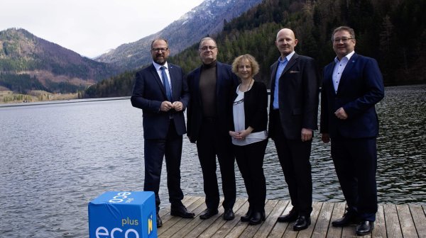 Fünf Personen stehen auf einem Holzsteg an einem ruhigen Bergsee, umgeben von bewaldeten Hängen und schneebedeckten Gipfeln.