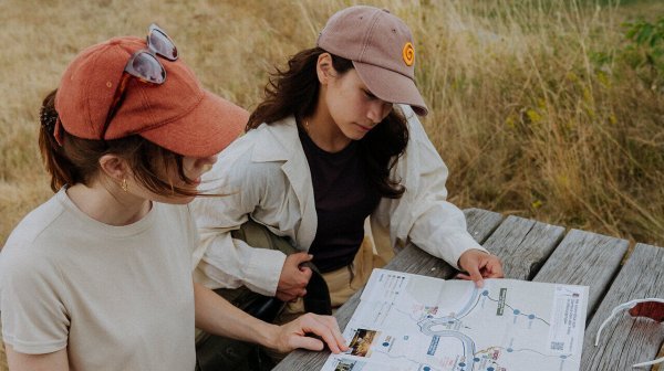 Zwei junge Frauen sitzen an einem Holztisch in einer naturnahen Landschaft und betrachten eine ausgebreitete Wander- oder Freizeitkarte. Eine weitere Person zeigt auf die Karte, während sie gemeinsam ihre Route planen.