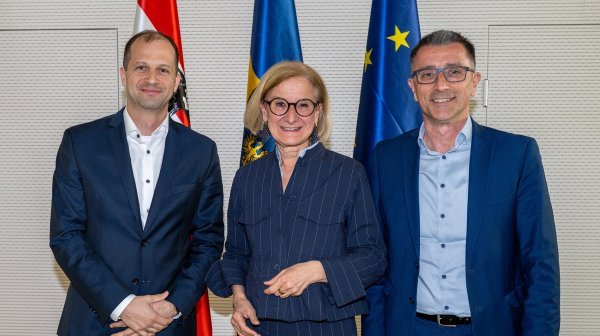 Drei erwachsene Personen stehen nebeneinander in einem Innenraum und blicken zur Kamera. Alle tragen formelle Kleidung. Hinter ihnen sind mehrere Flaggen sichtbar