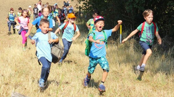 Eine große Gruppe von Kindern läuft auf einem trockenen, grasbewachsenen Weg im Freien. Im Vordergrund rennen mehrere Kinder mit Rucksäcken nebeneinander, einige tragen Kappen und bunte Kleidung. 