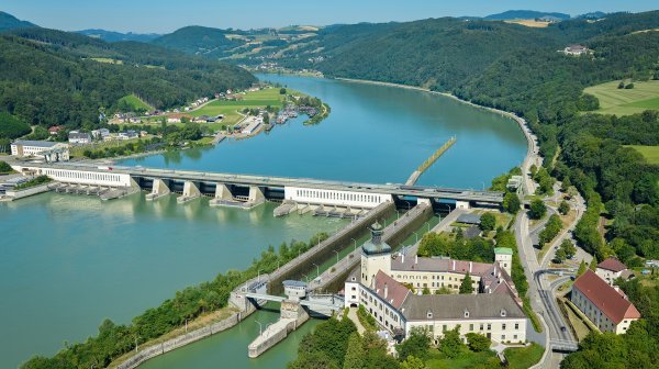 Luftaufnahme eines Wasserkraftwerks an einem breiten Fluss in einer bewaldeten Hügellandschaft. Im Vordergrund befinden sich Staumauer, Schleuse und ein historisches Gebäudekomplex, während sich der Fluss zwischen grünen Ufern und sanften Hügeln in die Ferne erstreckt.