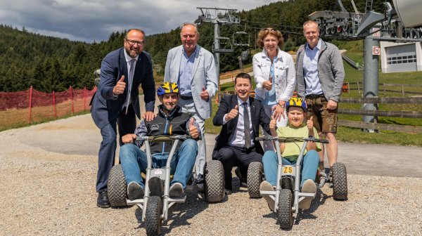 Gruppenfoto im Außenbereich einer Freizeitanlage in hügeliger Waldlandschaft. Zwei Personen sitzen mit Helmen auf dreirädrigen Mountaincarts im Vordergrund, während fünf weitere Personen dahinter und daneben stehen oder knien.