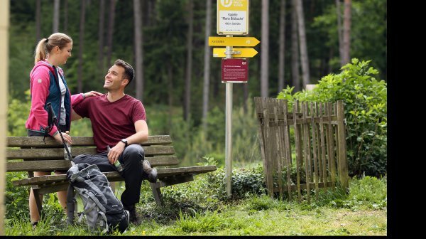 Zwei Wandernde machen eine Pause auf einer Holzbank am Waldrand. Ein Rucksack und Wanderstöcke stehen vor der Bank. Daneben befindet sich ein gelber Wegweiser der „Dunkelsteinerwaldrunde“ mit dem Hinweis zur Kapelle „Maria Bildbuche“. Im Hintergrund sind dichte Bäume und grüne Vegetation zu sehen.