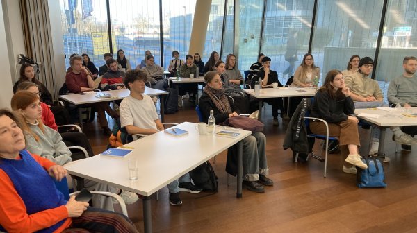 Personen sitzen bei Tischen und hören zu