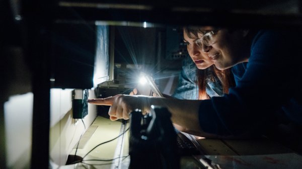Sehr dunkles Bild mit zwei Frauenprofilen und einem Lichtkegel auf ein Gerät 